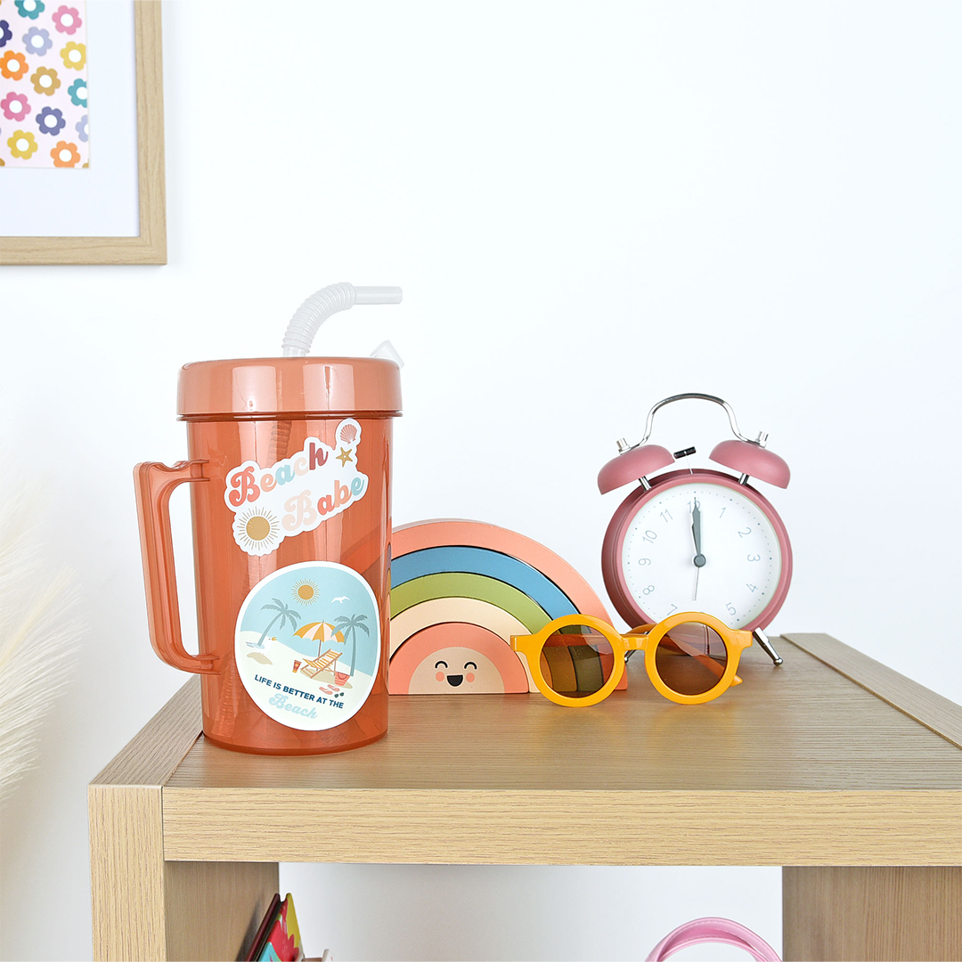 a water bottle sits on a night stand and has the "beach babe" sticker on it. the beach babe sticker says the words "beach babe" in colorful letters (pink, peach, teal) the words are surrounded by a sun, starfish, and seashell.