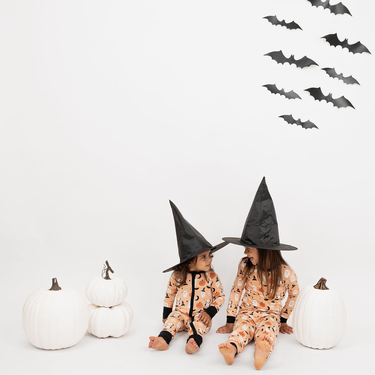 Kids wearing Boo-Tastic Halloween bamboo pajamas sitting with witch hats, white pumpkins, and bat wall backdrop.