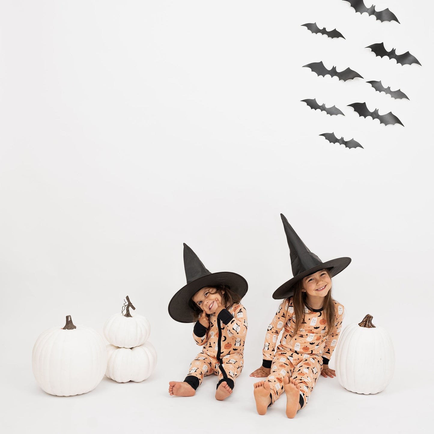 Two kids wearing Boo-Tastic Halloween bamboo pajamas with ghost and pumpkin print, smiling with witch hats and white pumpkins.