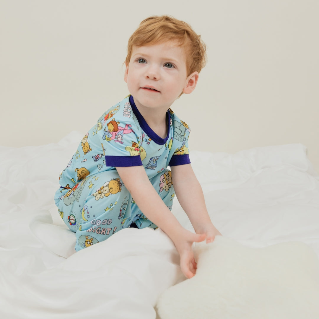 A toddler boy wearing our PBS Good Night Arthur™ collaboration, featuring pajamas adorned with Arthur and his friends in a cozy bedtime scene. The design includes storybooks, toothbrushes, bunny slippers, a sleeping puppy, crescent moons with sleep caps, twinkling stars, and the words 'GOOD NIGHT!' on a soft blue background.