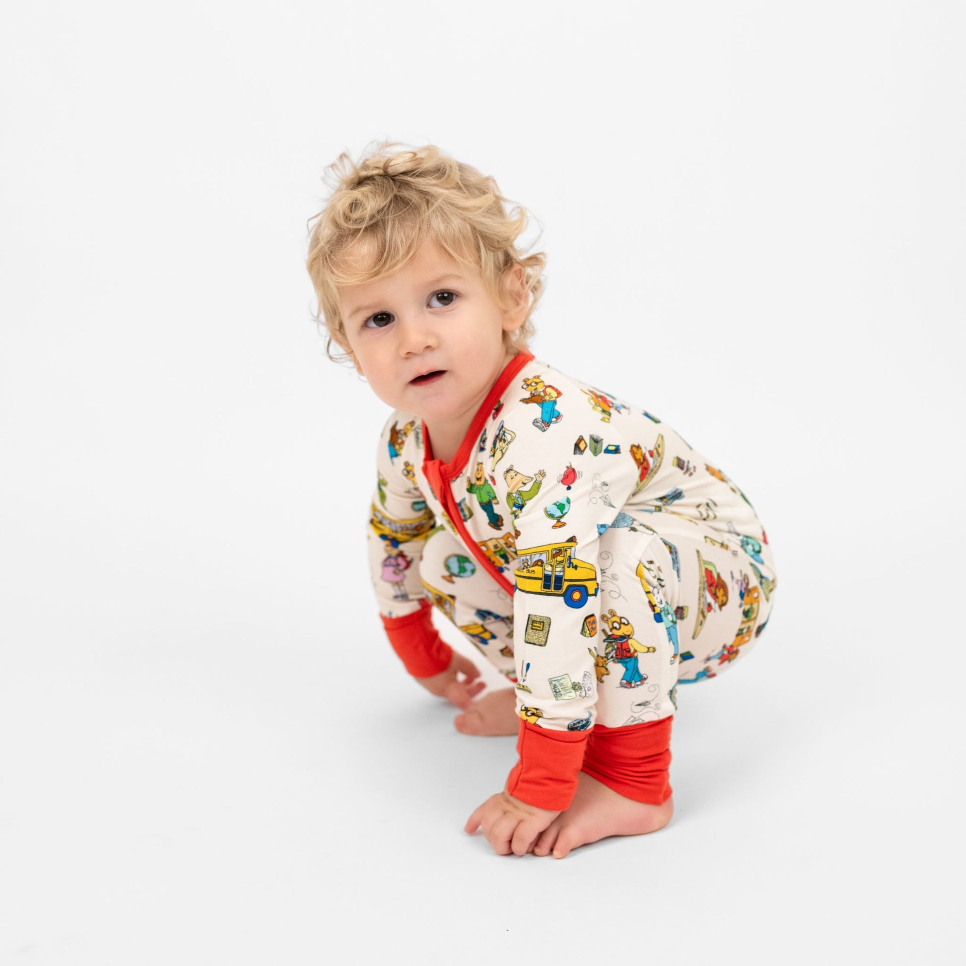 Baby boy squatting with  our buttery-soft  bamboo convertible pajama featuring red trims and playful illustrations of Arthur™ PBS Character, books, and school bus, a beige background.