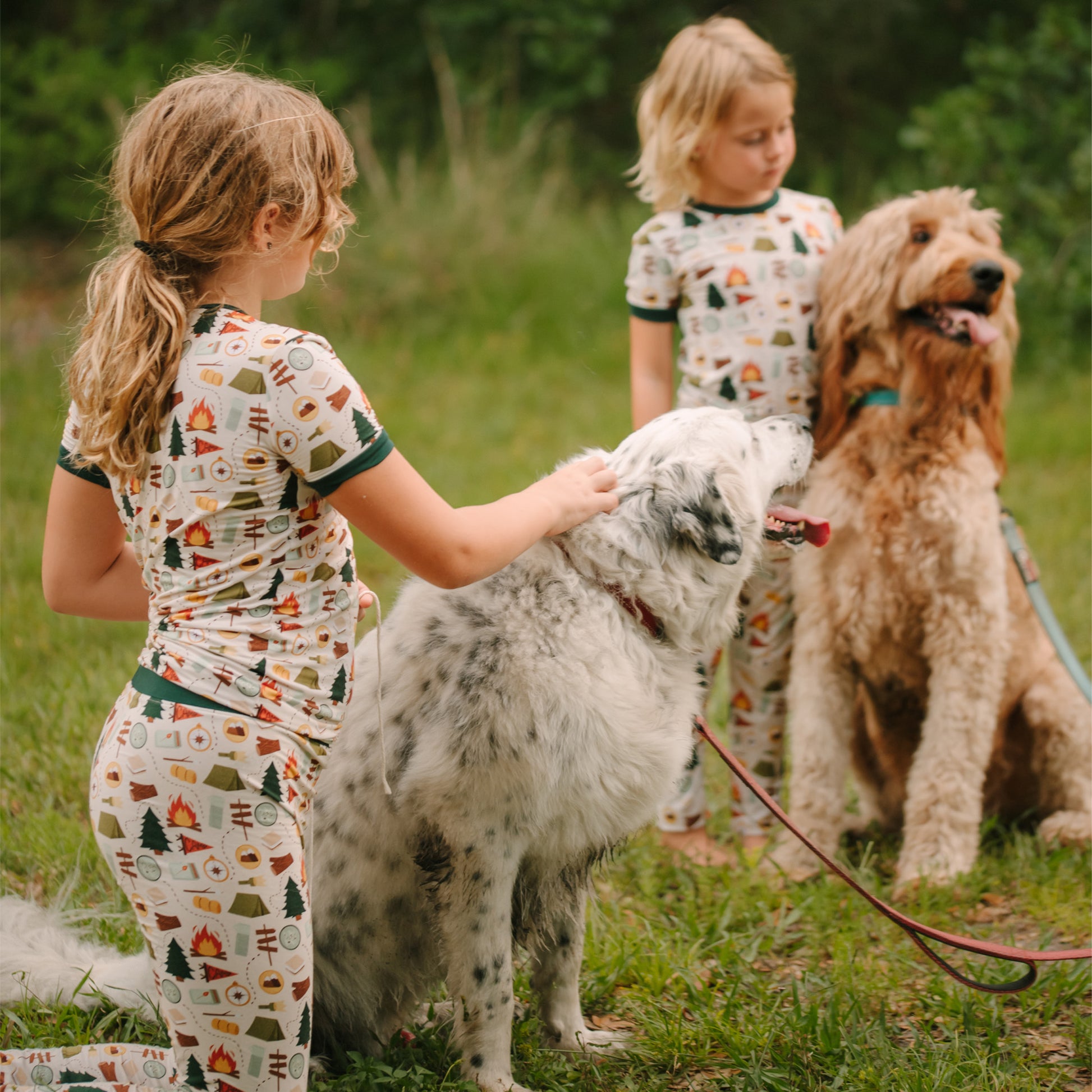 Young girls wearing camping print bamboo pajamas petting a fluffy white dog outdoors.