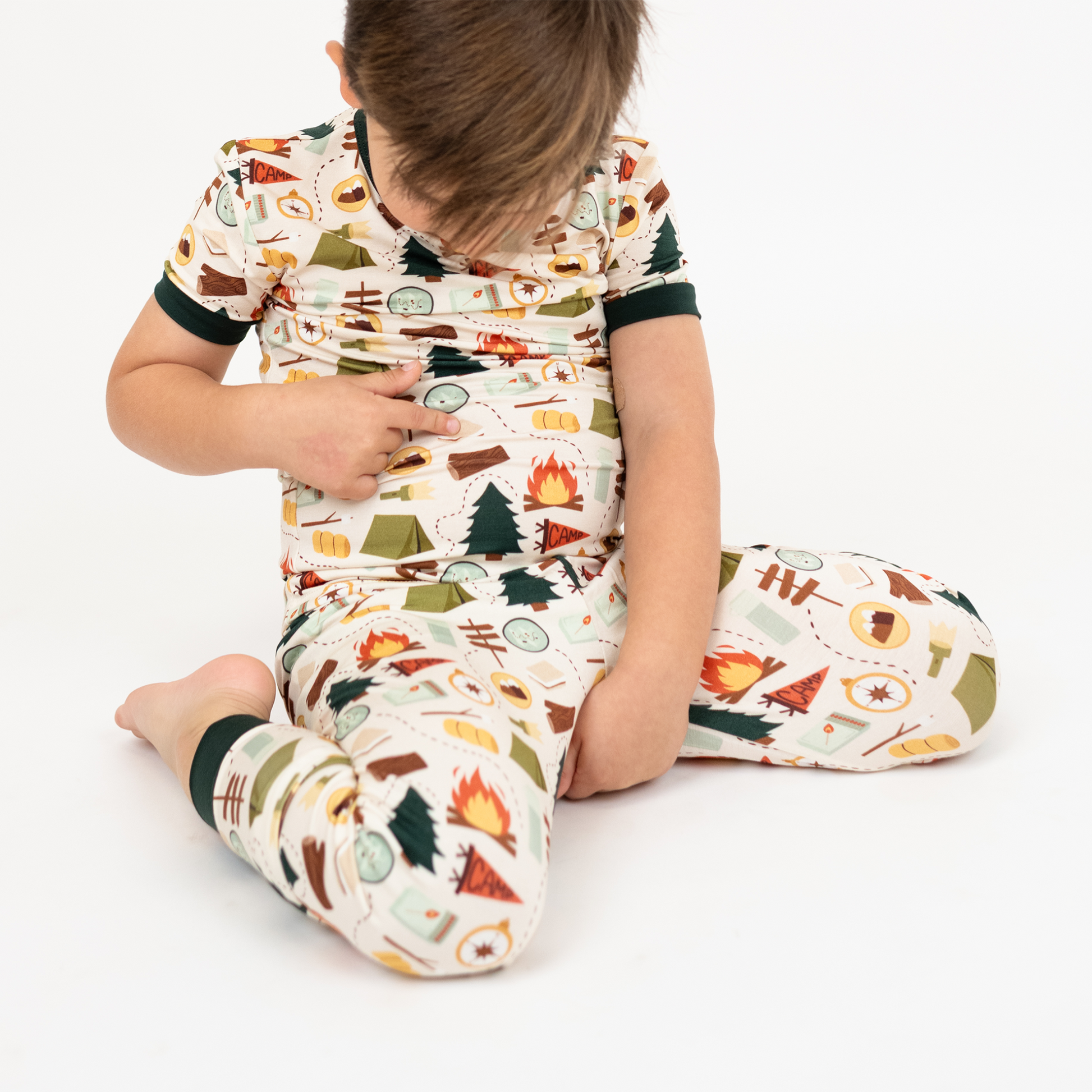 Boy sitting cross-legged on the floor wearing camping print bamboo pajamas.