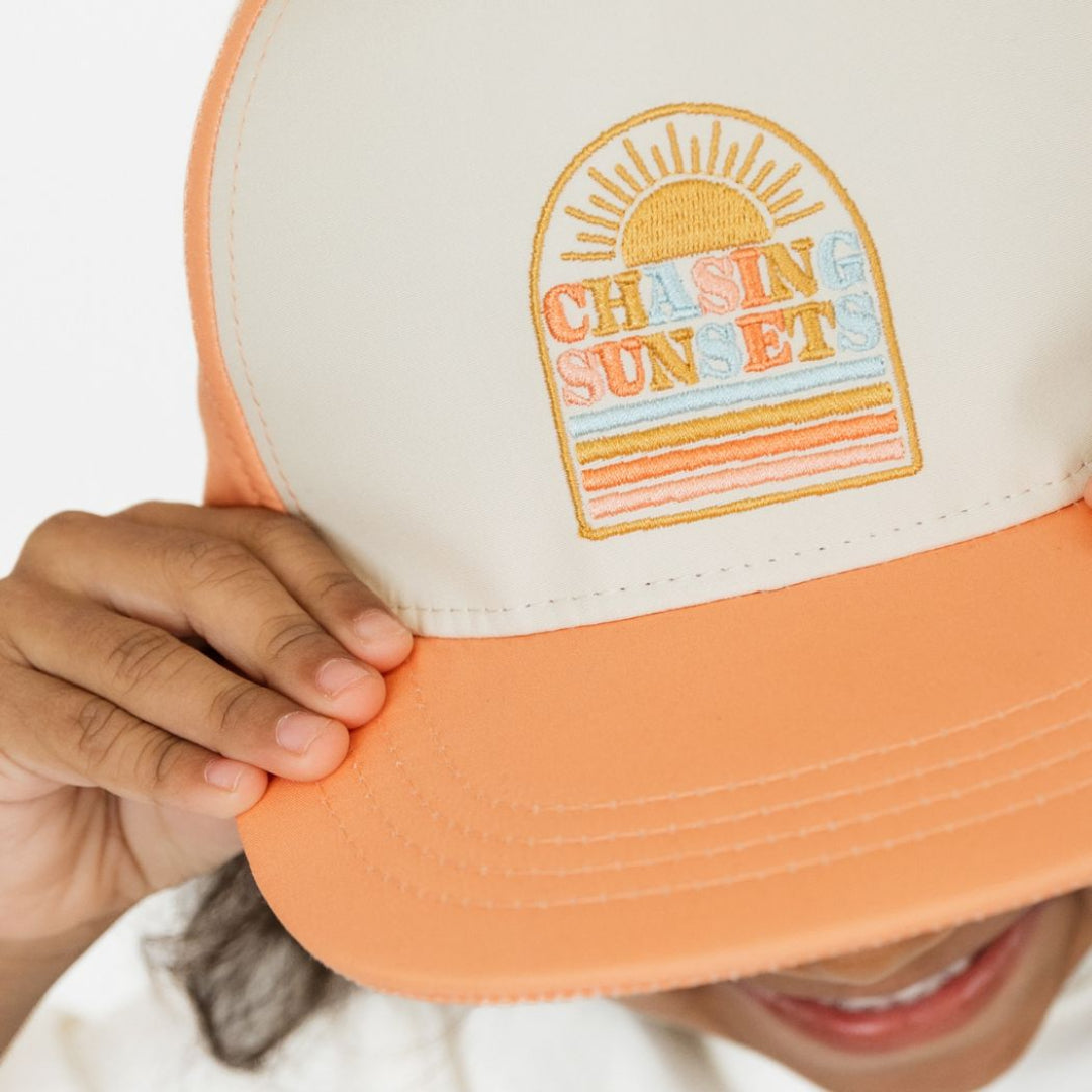 Little girl wearing our Chasing Sunsets Snapback hat holding onto the rim and smiling up at the camera.