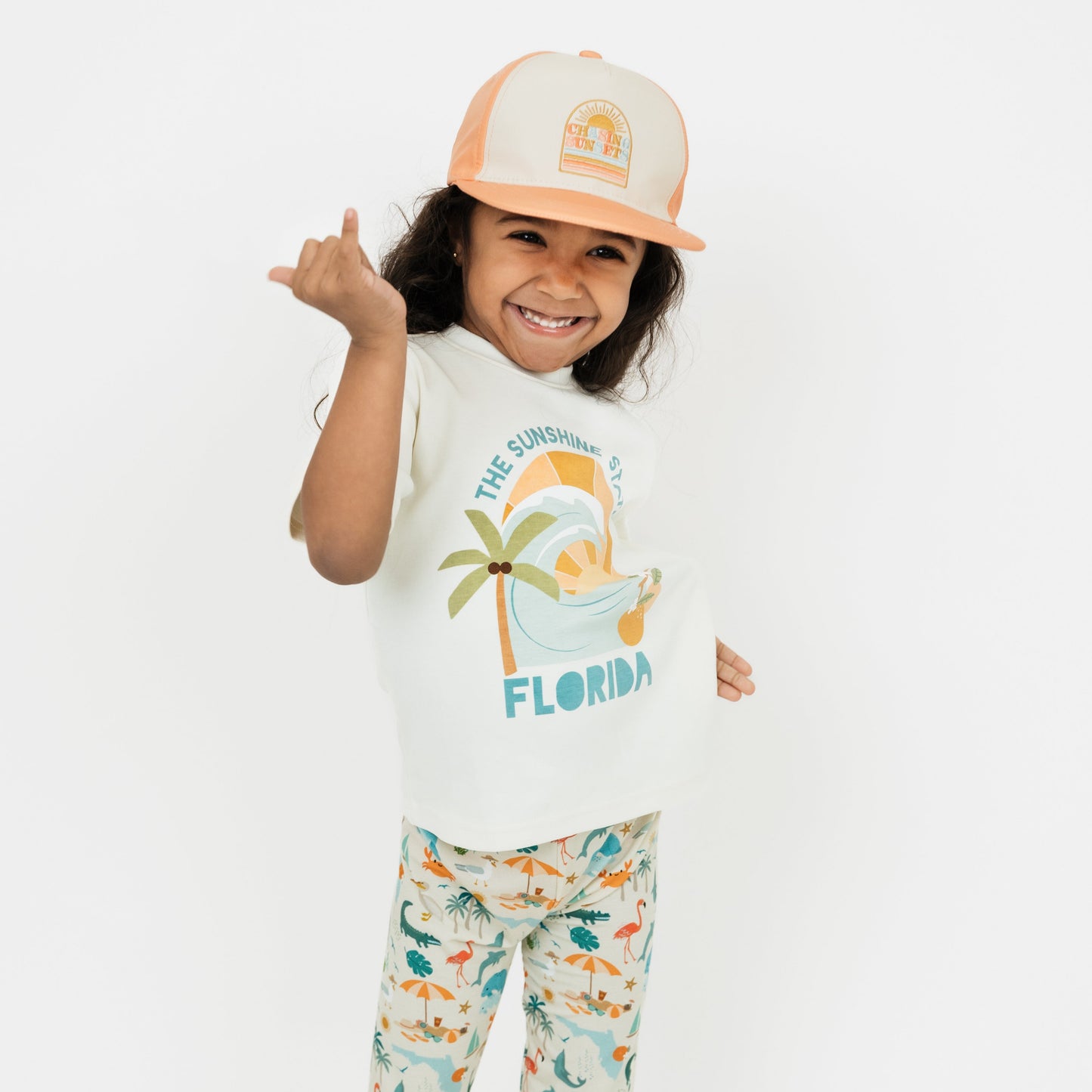 Little girl wearing our Chasing Sunsets Snapback Hat and The Sunshine State Florida Cotton Tee and Explore Florida Bamboo PJ pants holding up a shaka hand gesture.