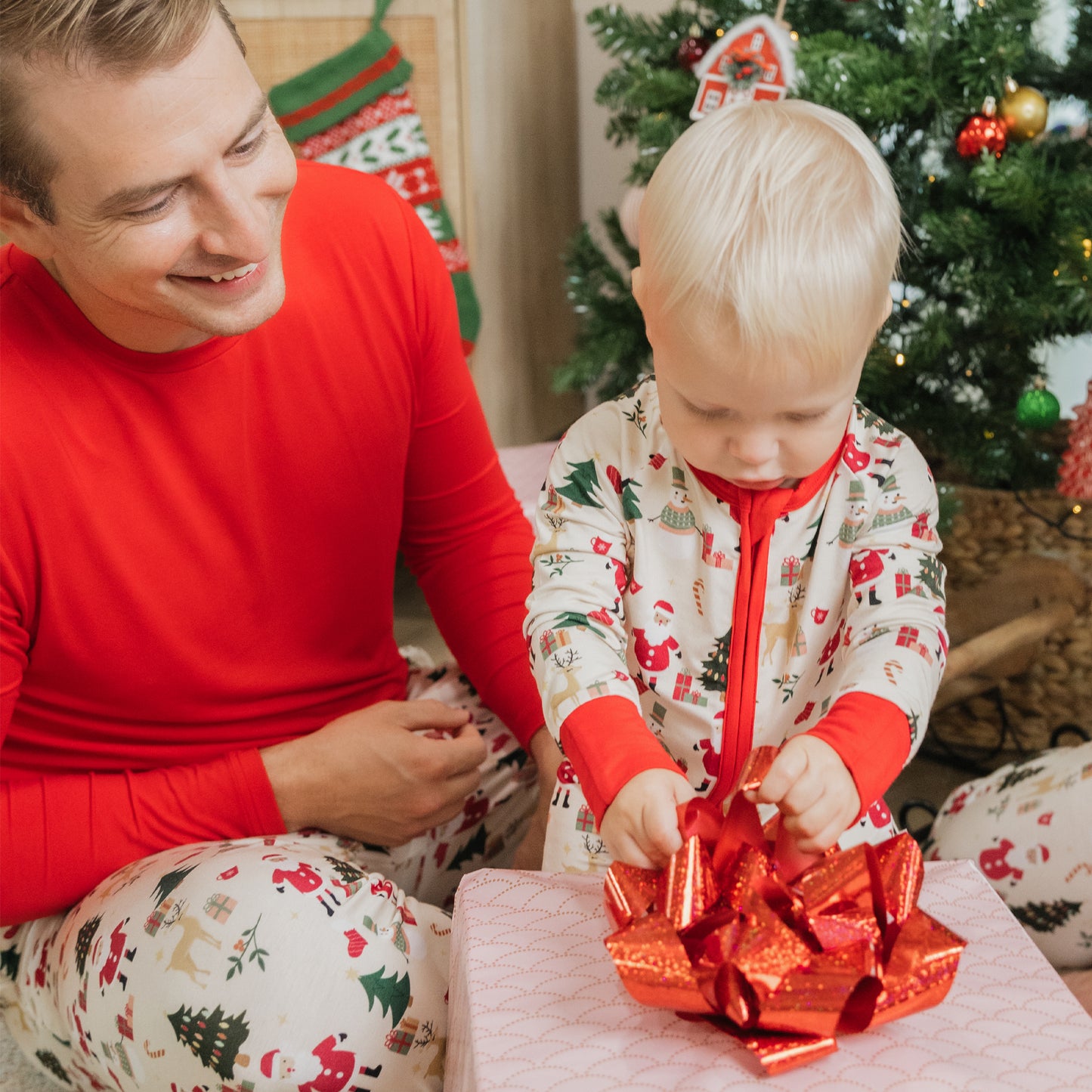 Parent and child wearing Christmas Red bamboo top and Cozy Christmas pajamas open presents together by the tree.
