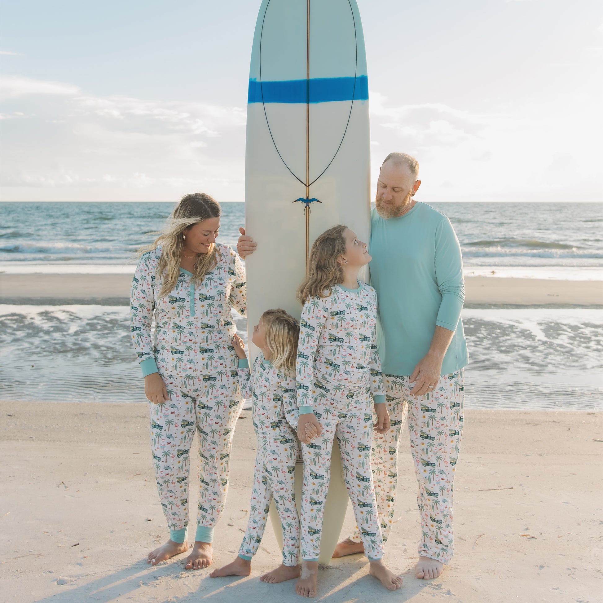 Family of four standing on a beach with a surfboard, all wearing matching pajamas.