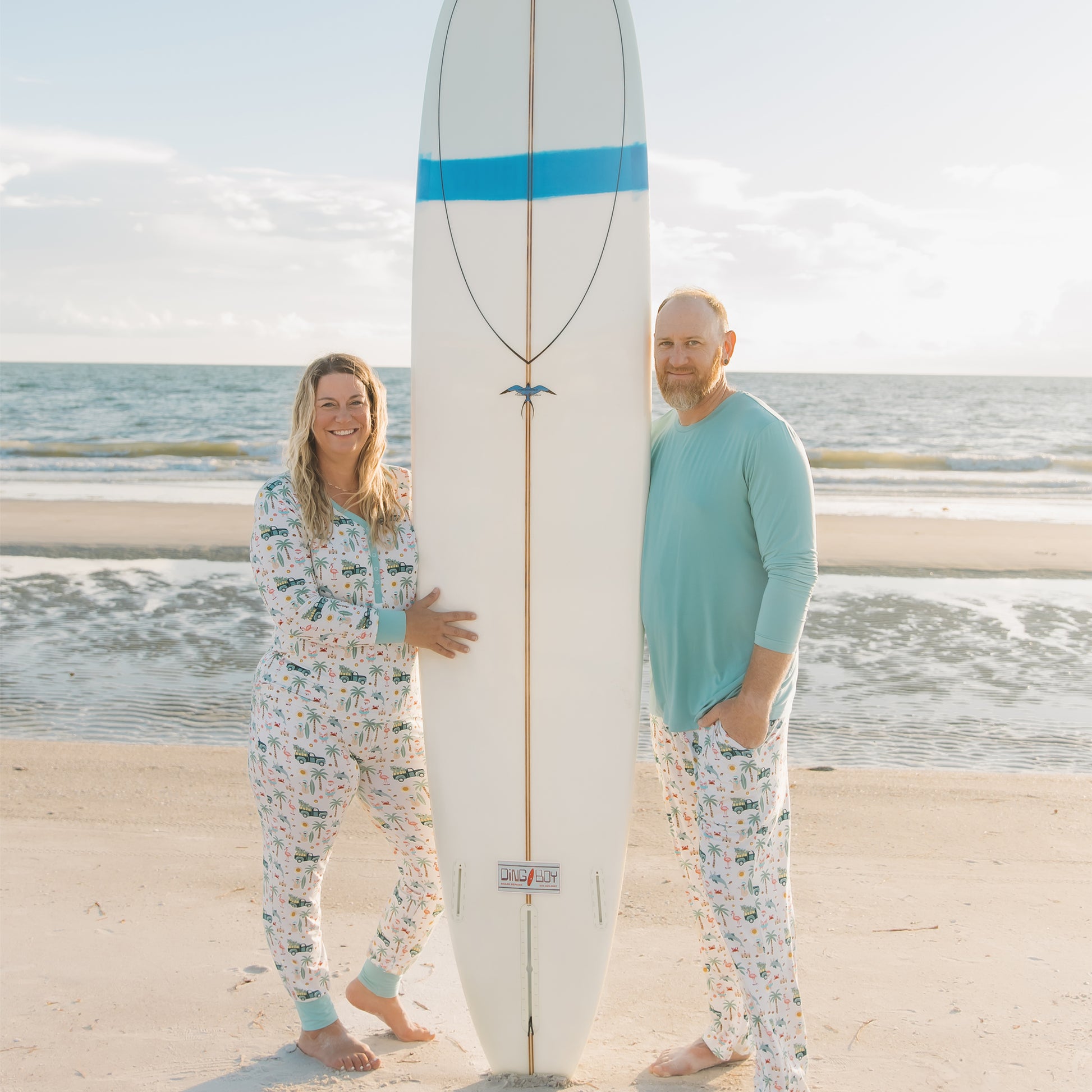 Two people in matching pajamas standing on a beach with a surfboard.