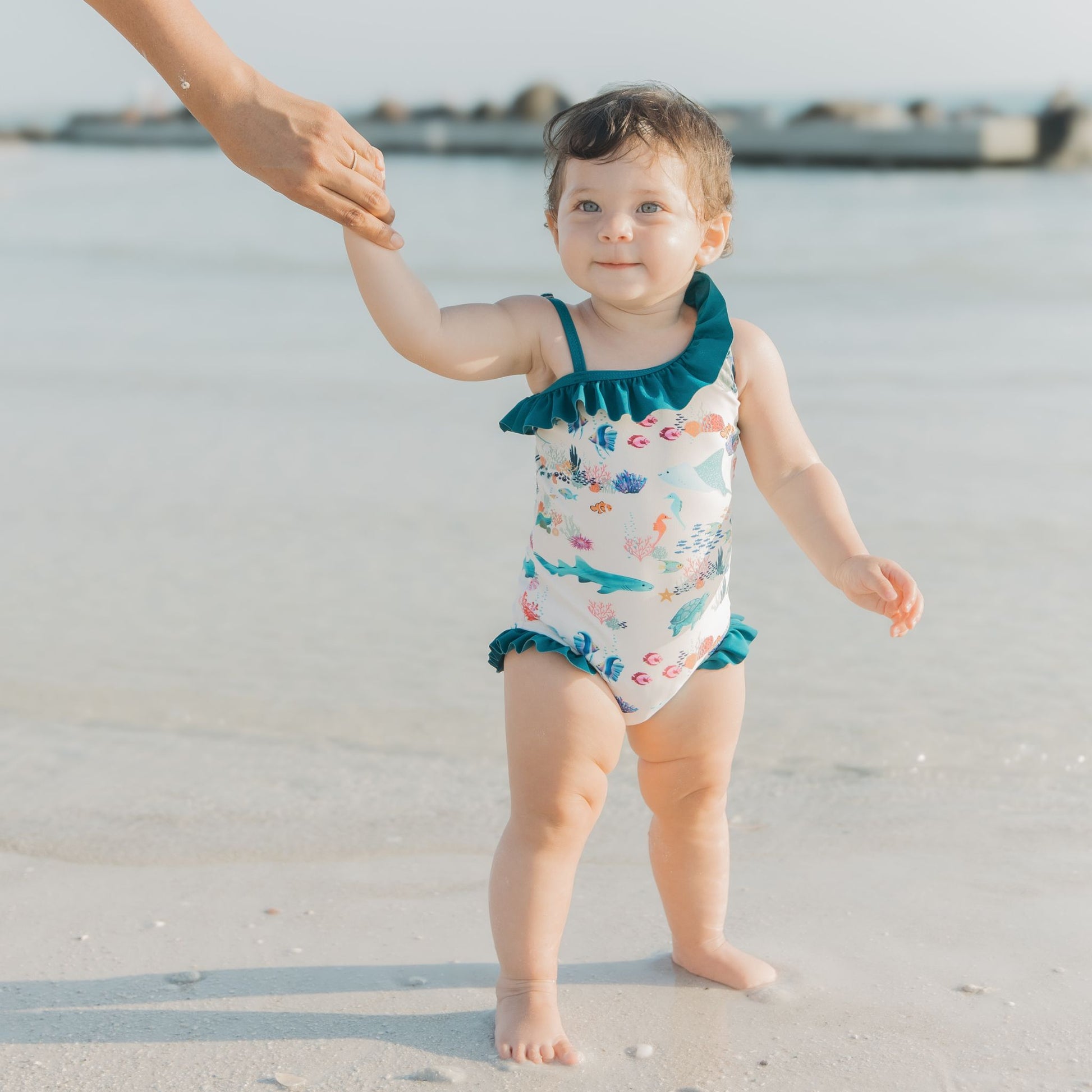Toddler girl wearing a Coral Reef Ruffle Leg One Piece Swimsuit with an ocean-themed print of sharks and sea turtles, designed with soft, stretchy fabric and UPF 50+ sun protection for a fun day at the beach.