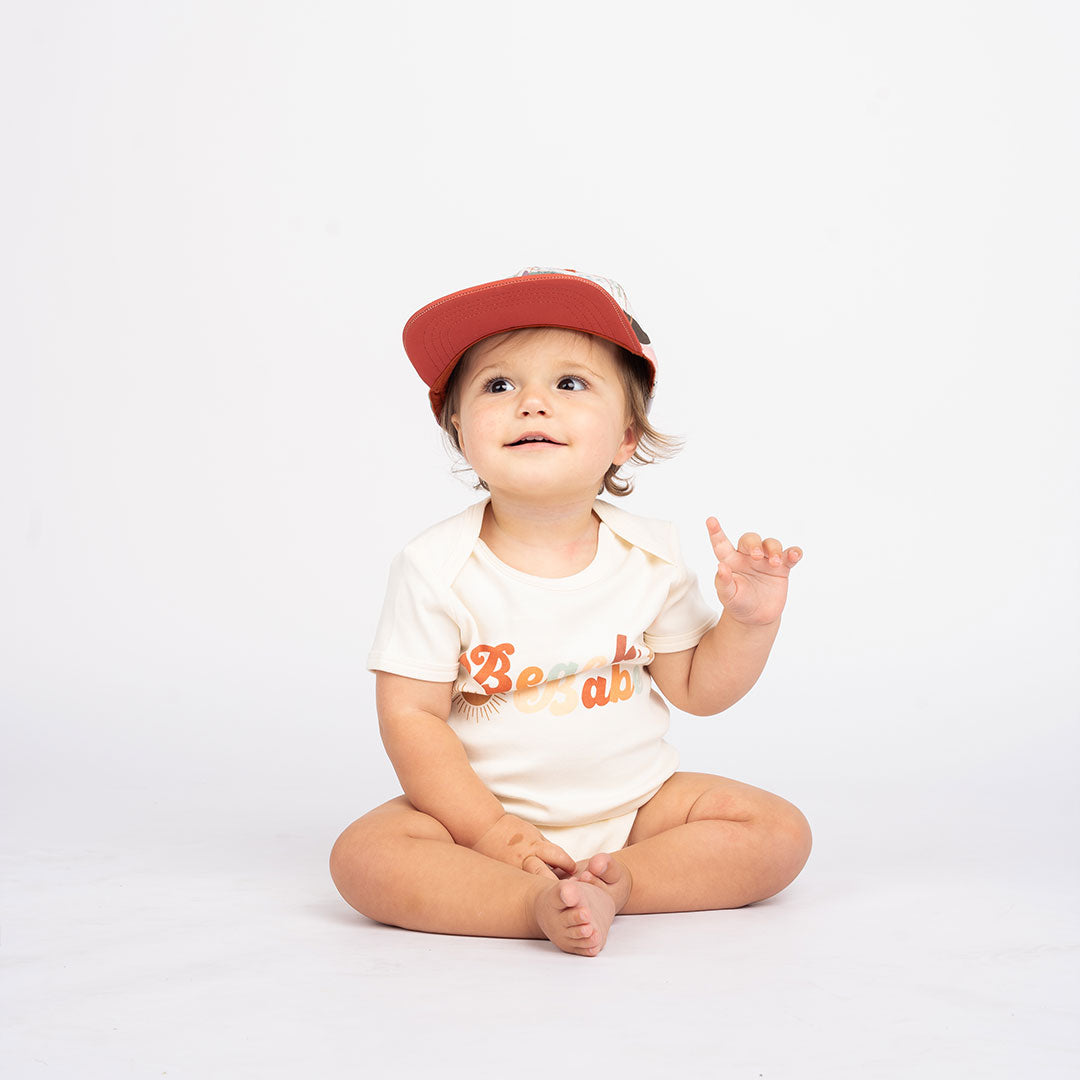 a baby sitting on the floor. she is wearing the "making waves" snap back hat and the the "beach babe" cotton bodysuit. the word 'beach babe' is spelt out in a groovy font with a sun, starfish, and shell surrounding it.