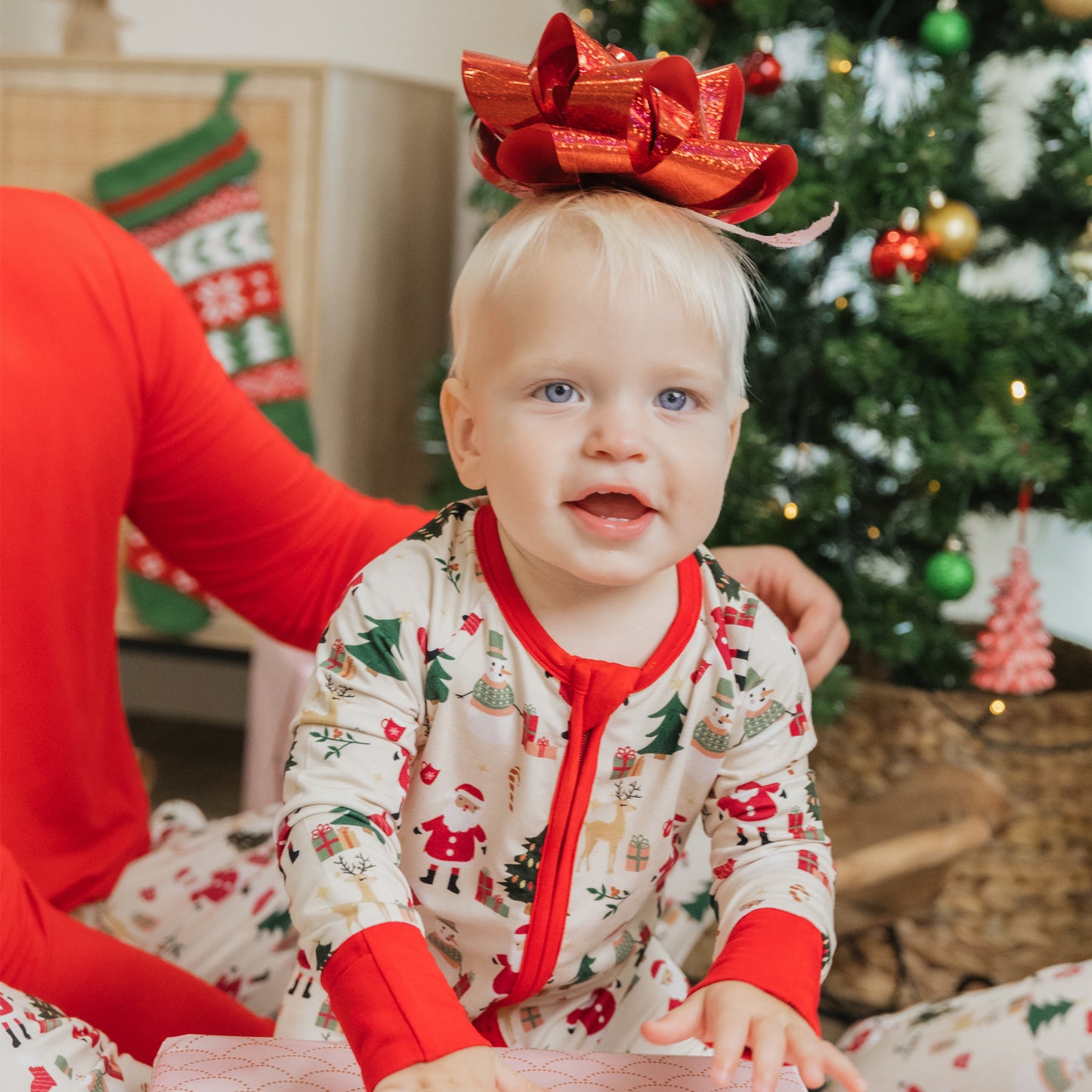 Baby wearing Cozy Christmas bamboo zipper pajama with red trim and holiday print, sitting under the tree with a festive bow.