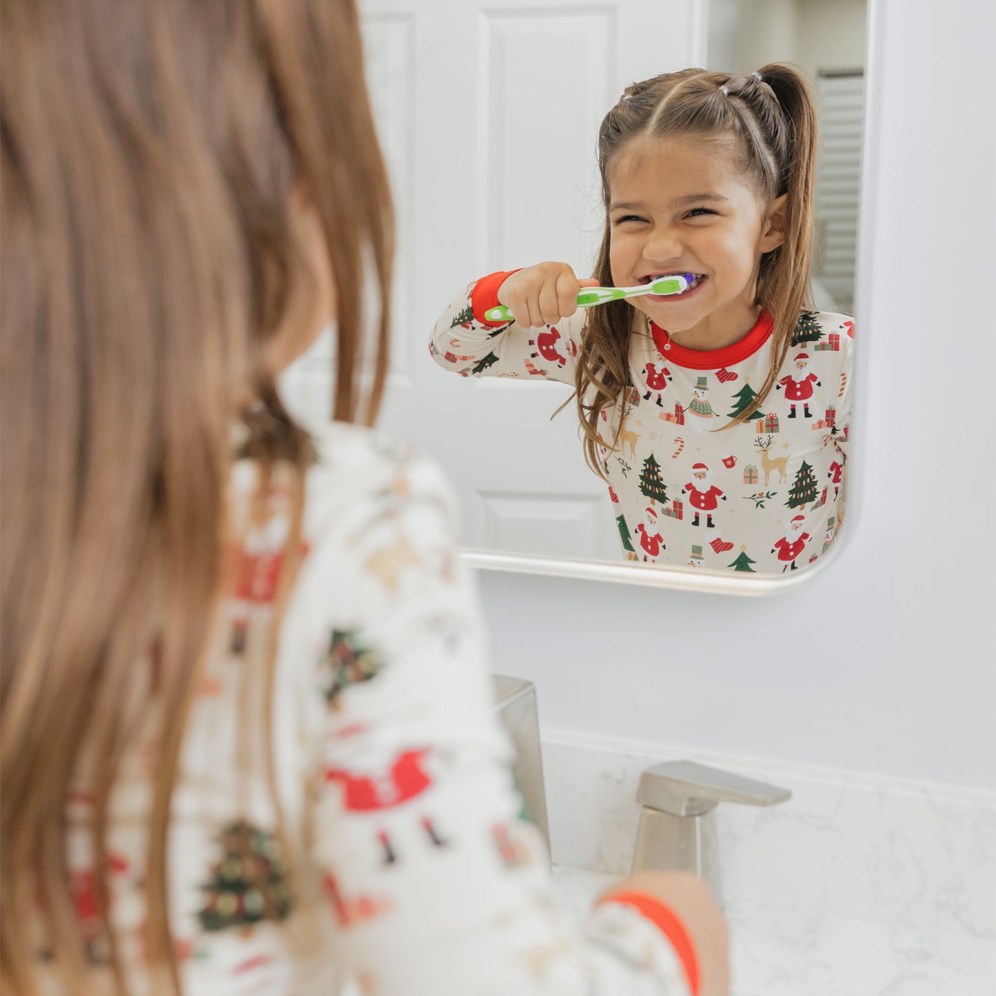 Girl wearing Cozy Christmas bamboo pajamas smiles in the mirror, showing off the festive holiday print with red trim.
