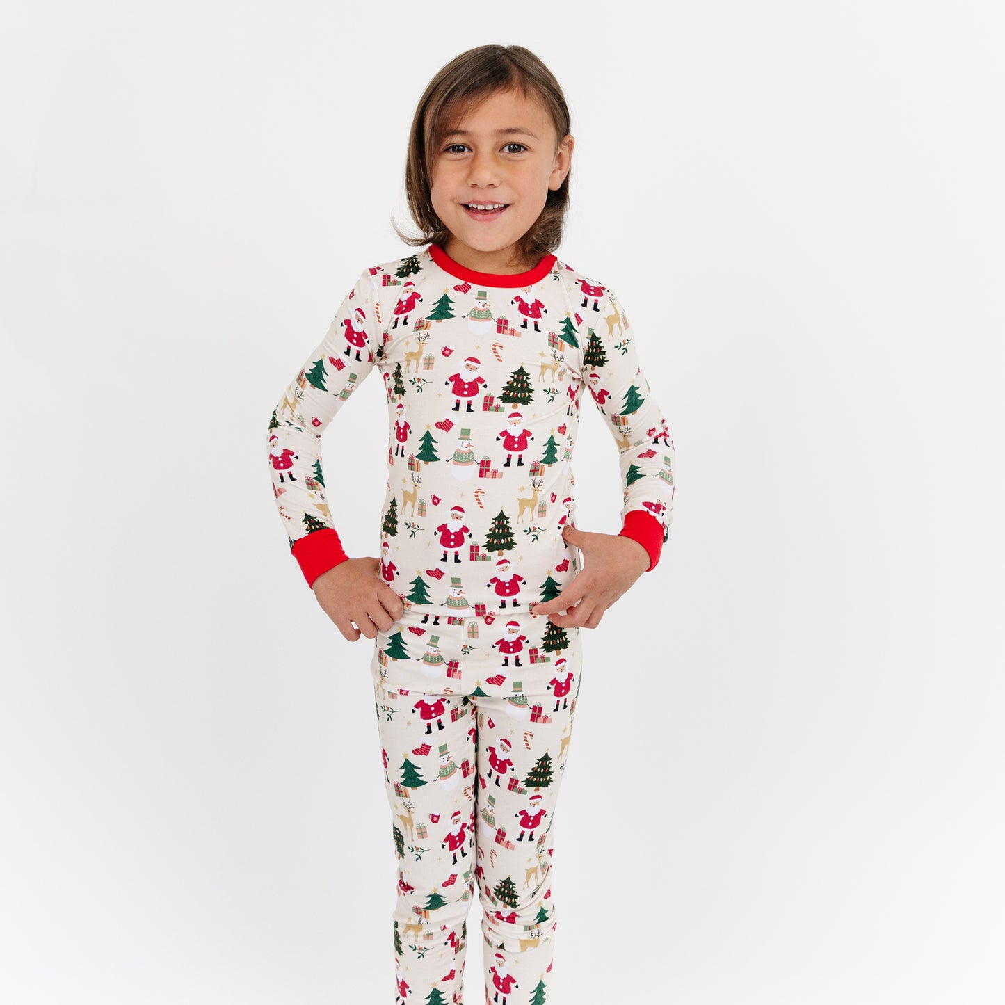 Child standing in studio wearing Cozy Christmas bamboo pajamas, featuring Santa and tree print with bright red cuffs.