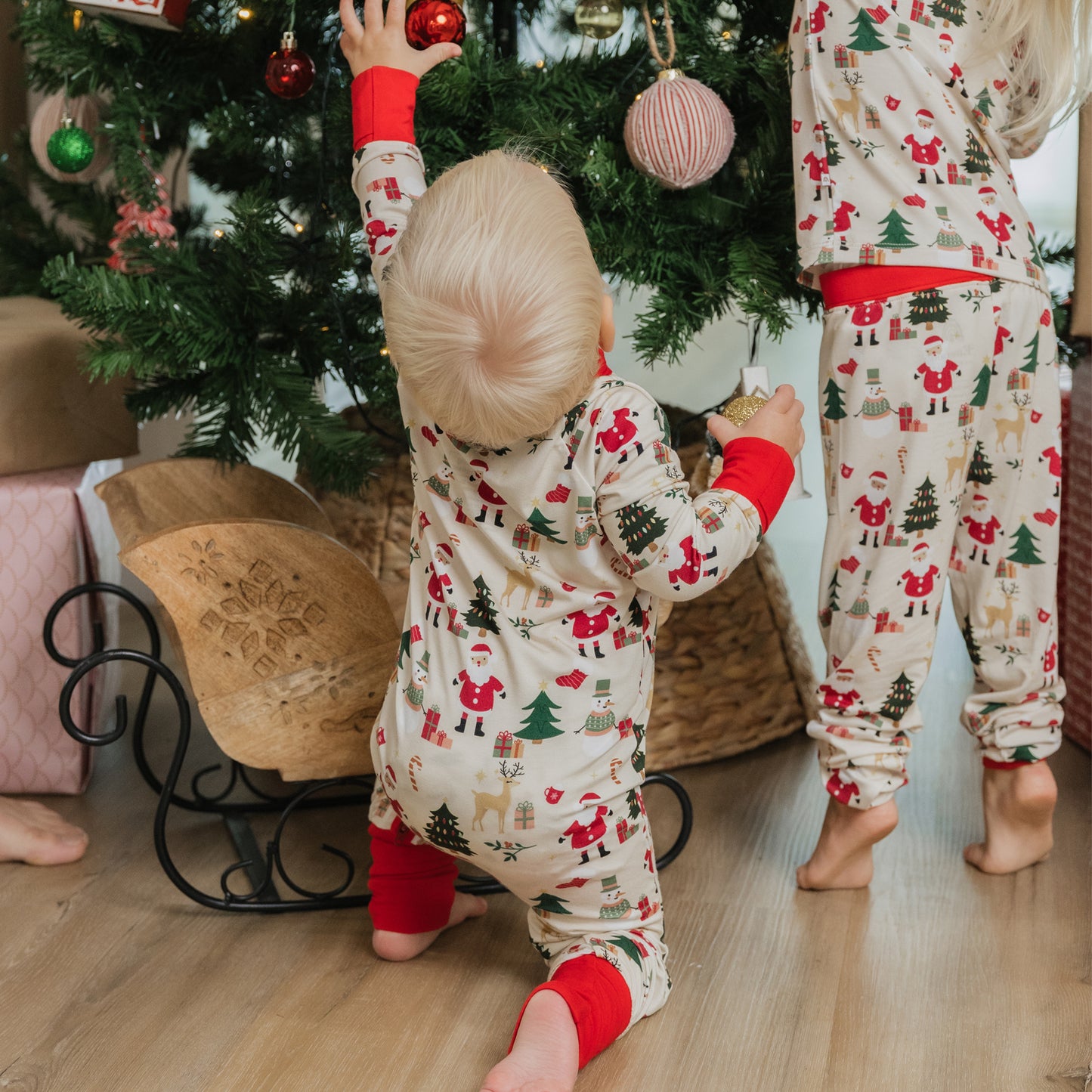 Two toddlers wearing Cozy Christmas bamboo zip pajamas decorating a Christmas tree – festive holiday sleepwear for babies and kids.