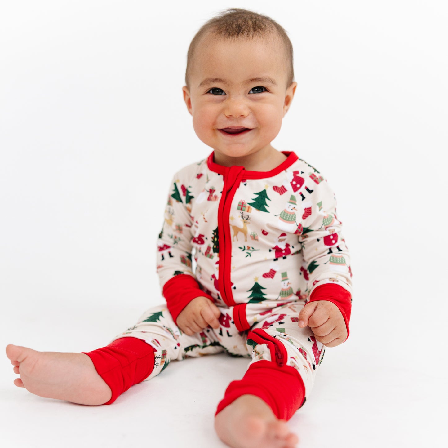 Smiling baby sitting in Cozy Christmas bamboo zipper pajamas with Santa and reindeer print – perfect for cozy holiday mornings.
