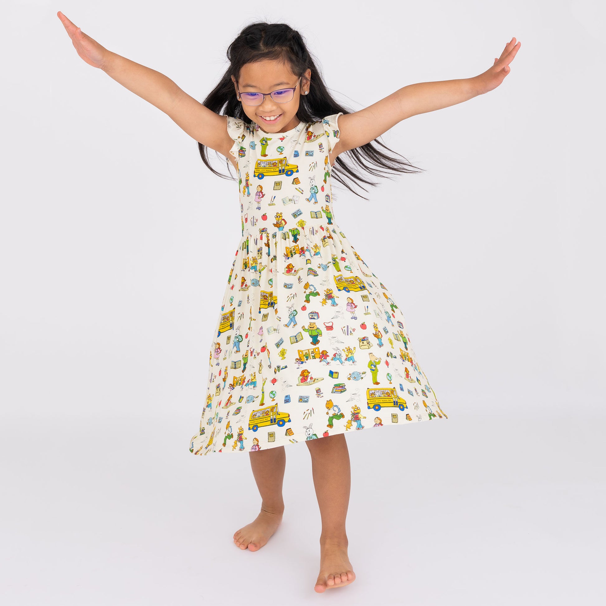 A happy girl twirls in our bamboo twirl dress, featuring playful illustrations of Arthur™ from PBS, books, and a school bus on a soft beige background.