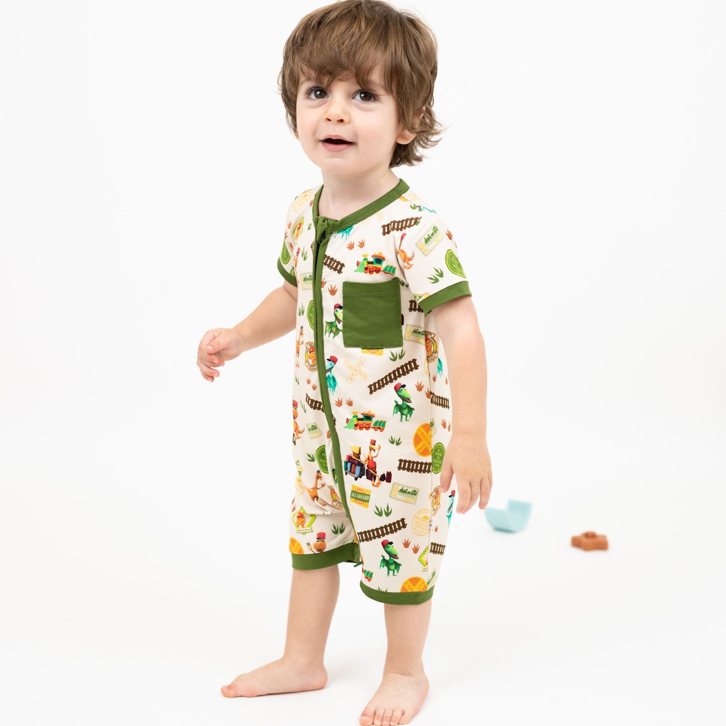 Toddler wearing our Emerson and Friends All Aboard the Dinosaur Train™ Bamboo Baby Shortie Romper featuring with green trim, and colorful orange, blue, and green dinosaurs on train tracks with "Dino Train" print.