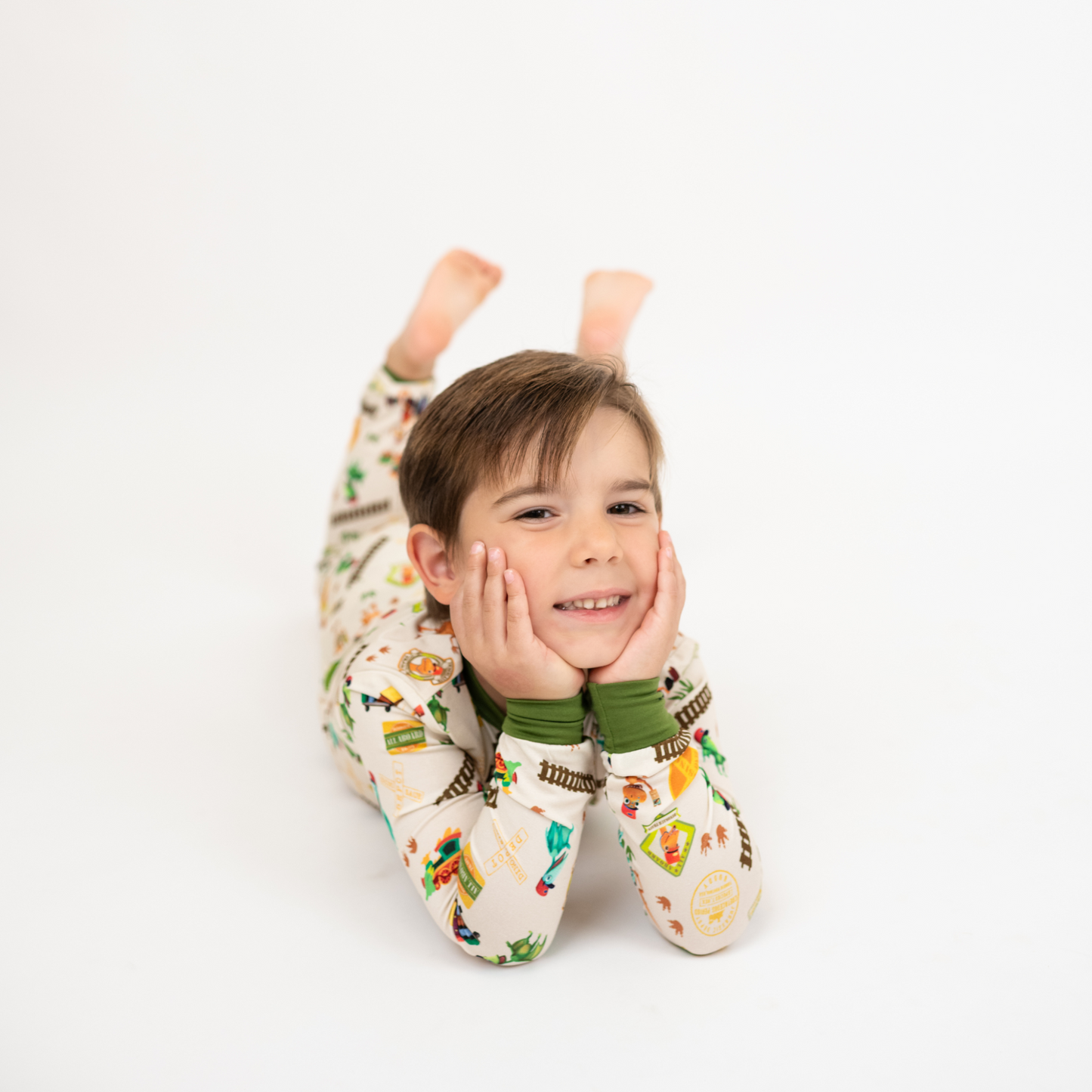 Toddler wearing our Emerson and Friends All Aboard the Dino Train Long Sleeve Kids Pajama Pants Set with green trim, and colorful orange, blue, and green dinosaurs on train tracks with "Dino Train" print.