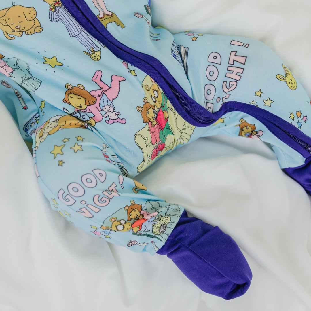 A close-up of the double zipper convertible pajama from our PBS Good Night Arthur™ collaboration, featuring a cozy bedtime scene with Arthur and friends, storybooks, toothbrushes, bunny slippers, a sleeping puppy, crescent moons with sleep caps, twinkling stars, and the words "GOOD NIGHT!" on a soft blue background.