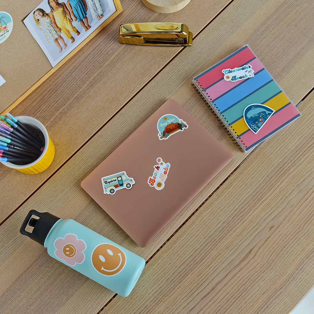 the "explore camper van" sticker on a brown laptop. the "explore camper van" sticker. a blue and white van with orange tones. the word "explore" is printed on the window. you can also see the "beach babe" sticker on the laptop too. next to the laptop you can see the "clearwater beach" and "dolphin" sticker on a rainbow notebook.