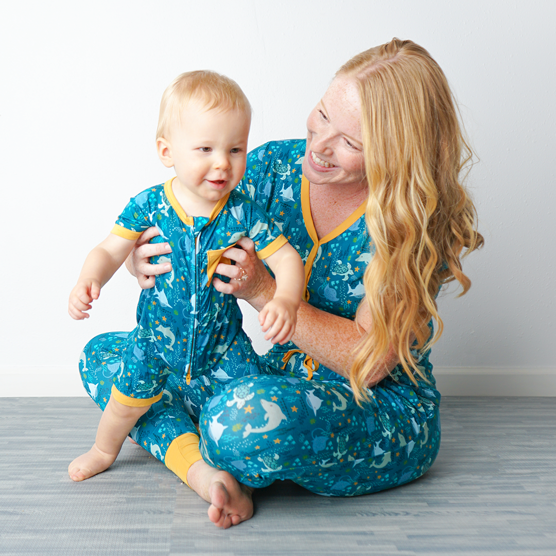 mom sits criss-cross on the floor with her baby in her hands. the baby stands in his "ocean friends" shortie. the woman wears the "ocean friends" women's top and jogger bottoms. the "ocean friends" print is a combination of dolphins, stingrays, fish, starfish, coral, bubbles, and sharks, all spread out on a deep sea blue background.