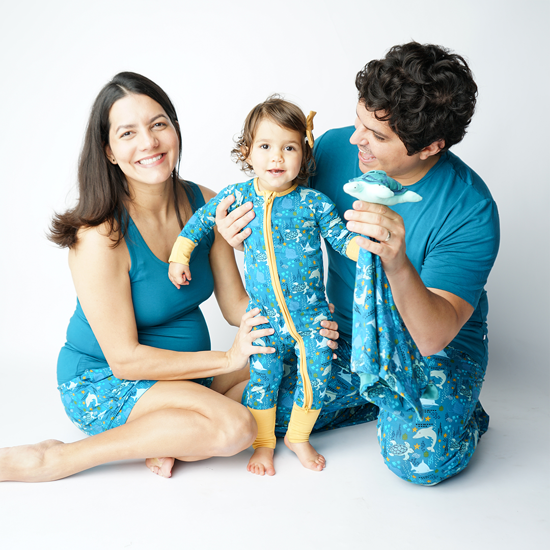 a family of 3 sit on the floor, all wearing different versions of the "ocean friends prints" the mom smiles in the women's shorts and holds on to her baby in the convertible. the dad smiles at his daughter, while wearing the relaxed pants. they both hold the lovey. the "ocean friends" print is a combination of dolphins, stingrays, fish, starfish, coral, bubbles, and sharks, all spread out on a deep sea blue background.