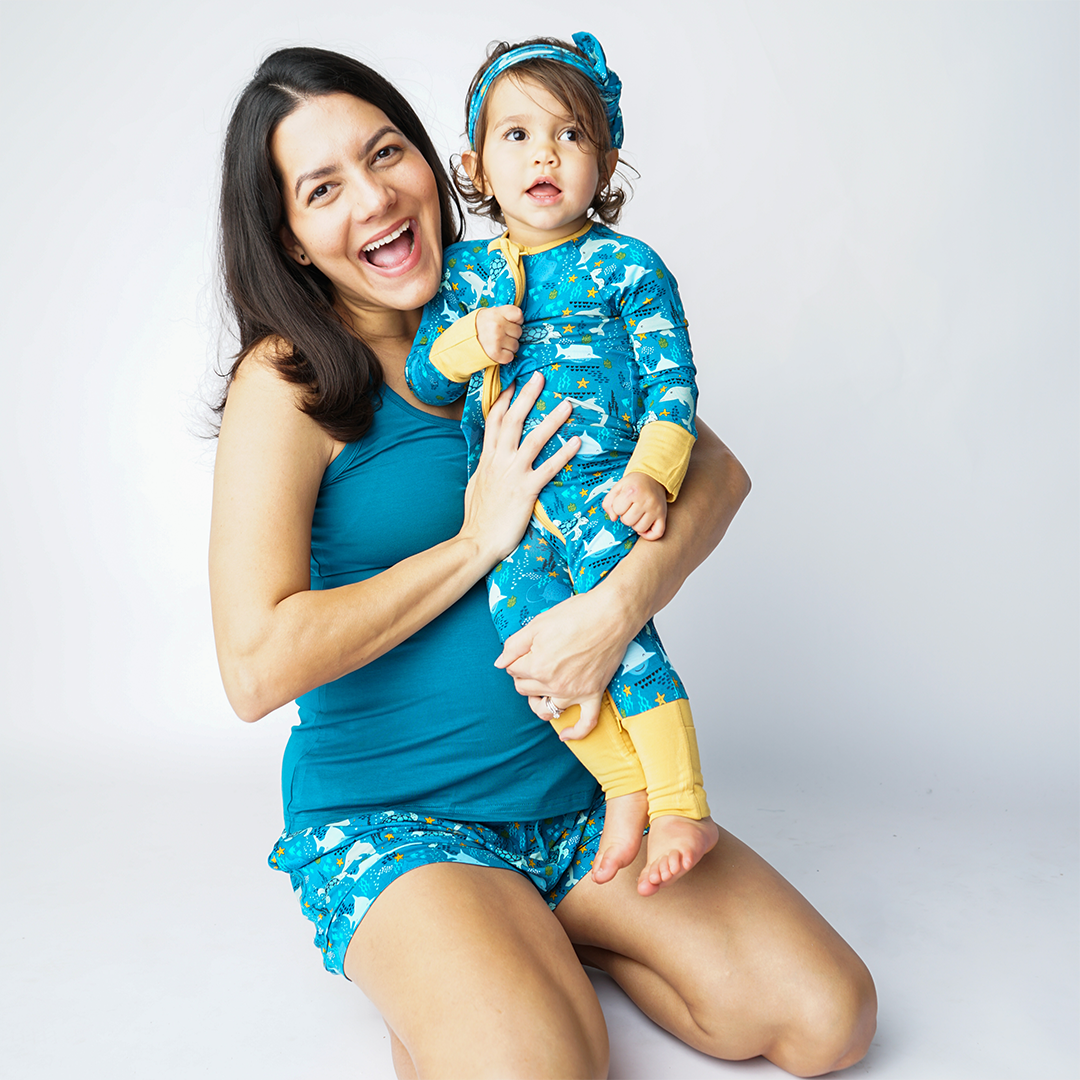 mom smiles while sitting on the floor.  with her baby standing in her lap. the mom wears the "ocean friends" women's shorts and the "ocean blue" tank top. the baby wears the "ocean friends" convertible. the "ocean friends" print is a combination of dolphins, stingrays, fish, starfish, coral, bubbles, and sharks, all spread out on a deep sea blue background.