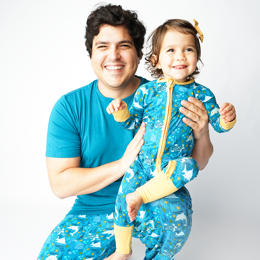 dad and daughter smile together. the dad sits on his knees in the "ocean friends" relaxed pants and the "ocean blue" unisex top. he hold his baby girl in her "ocean friends" convertible. the "ocean friends" print is a combination of dolphins, stingrays, fish, starfish, coral, bubbles, and sharks, all spread out on a deep sea blue background.