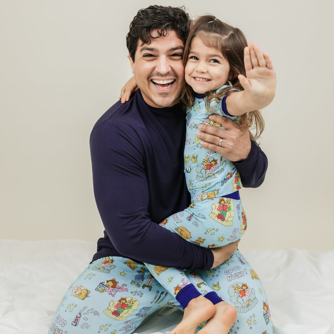 A heartwarming moment captures the father with daughter smiling, with them wearing our PBS Good Night Arthur™ collaboration featuring pajamas adorned with Arthur and friends in a cozy bedtime scene complete with storybooks, toothbrushes, bunny slippers, a sleeping puppy, crescent moons with sleep caps, twinkling stars, and the words "GOOD NIGHT!" on a soft blue background.