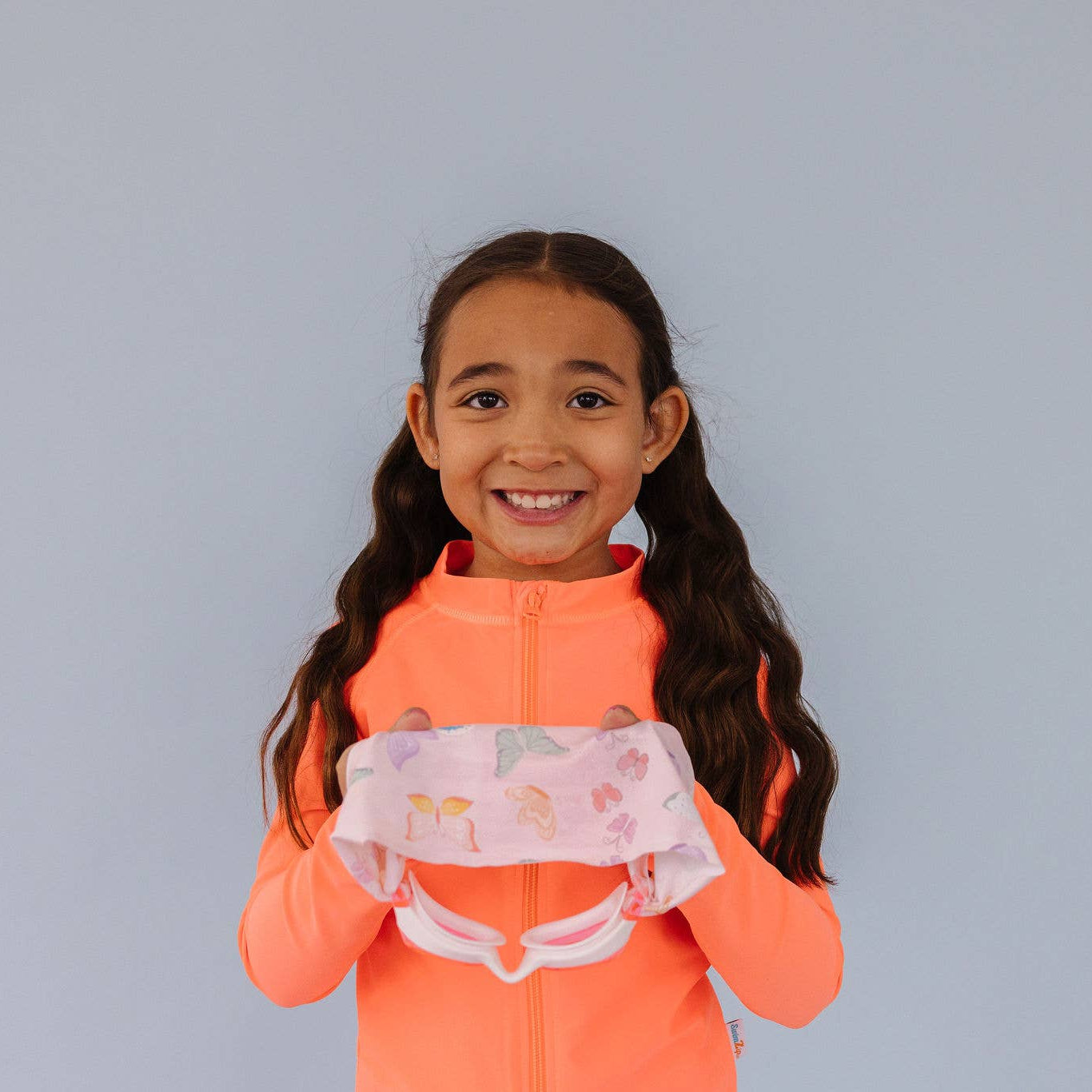 Young girl in an orange jacket holding Splash Place flutter swim goggles with butterfly print fabric strap against a gray background