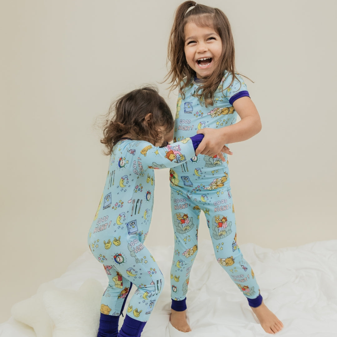 A heartwarming moment captures two sisters smiling, with them wearing our PBS Good Night Arthur™ collaboration featuring pajamas adorned with Arthur and friends in a cozy bedtime scene complete with storybooks, toothbrushes, bunny slippers, a sleeping puppy, crescent moons with sleep caps, twinkling stars, and the words "GOOD NIGHT!" on a soft blue background.