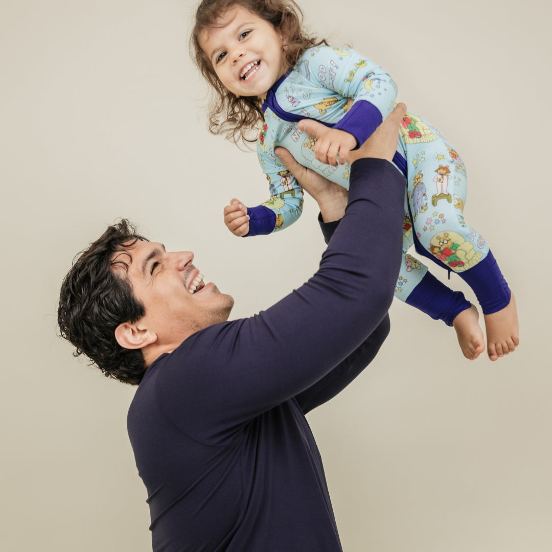A heartwarming moment captures a dad and his smiling daughter, with her wearing our PBS Good Night Arthur™ collaboration featuring pajamas adorned with Arthur and friends in a cozy bedtime scene complete with storybooks, toothbrushes, bunny slippers, a sleeping puppy, crescent moons with sleep caps, twinkling stars, and the words "GOOD NIGHT!" on a soft blue background.