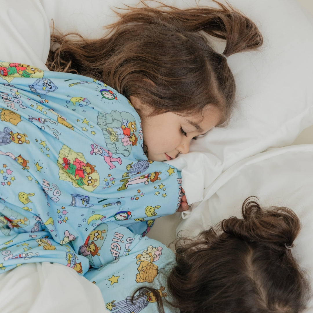 Sisters laying down sleeping with our blanket and our PBS Good Night Arthur™ collaboration featuring pajamas adorned with Arthur and friends in a cozy bedtime scene complete with storybooks, toothbrushes, bunny slippers, a sleeping puppy, crescent moons with sleep caps, twinkling stars, and the words "GOOD NIGHT!" on a soft blue background.