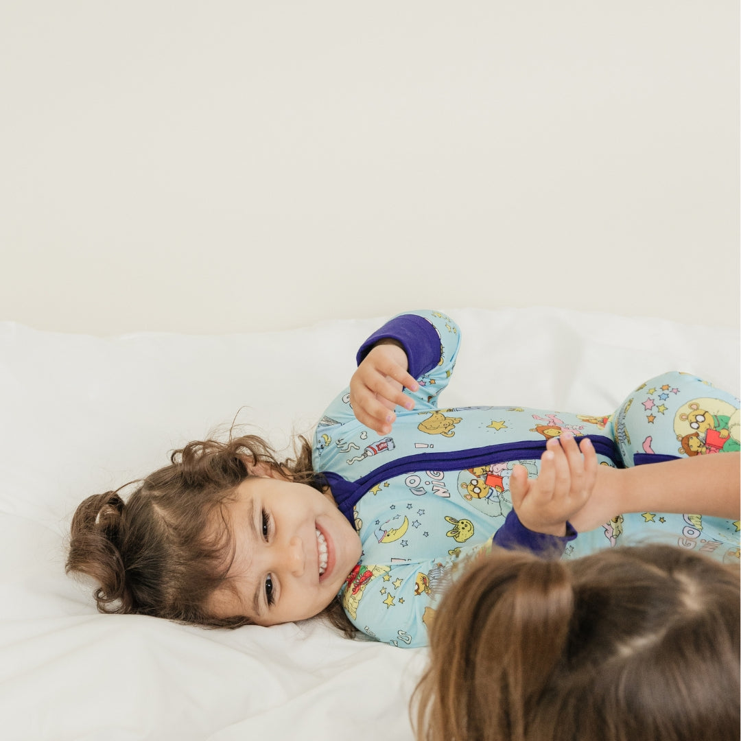 Adorable moment with sisters smiling. She is wearing our collaboration with PBS Good Night Arthur™ pattern. The pajama features Arthur and friends in a cozy bedtime scene with storybooks, toothbrushes, bunny slippers, a sleeping puppy, crescent moons wearing sleep caps, twinkling stars, and the words "GOOD NIGHT!" on a soft blue background.