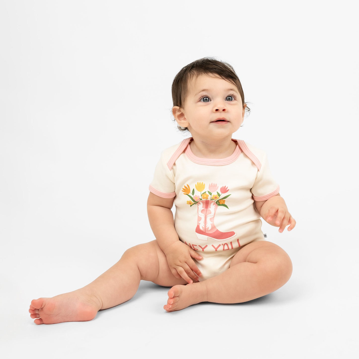 A cute baby wearing our Hey Y'all onesie paired with a pink cowgirl boots print and floral accents, capturing the essence of rodeo and western style for a perfect outfit.