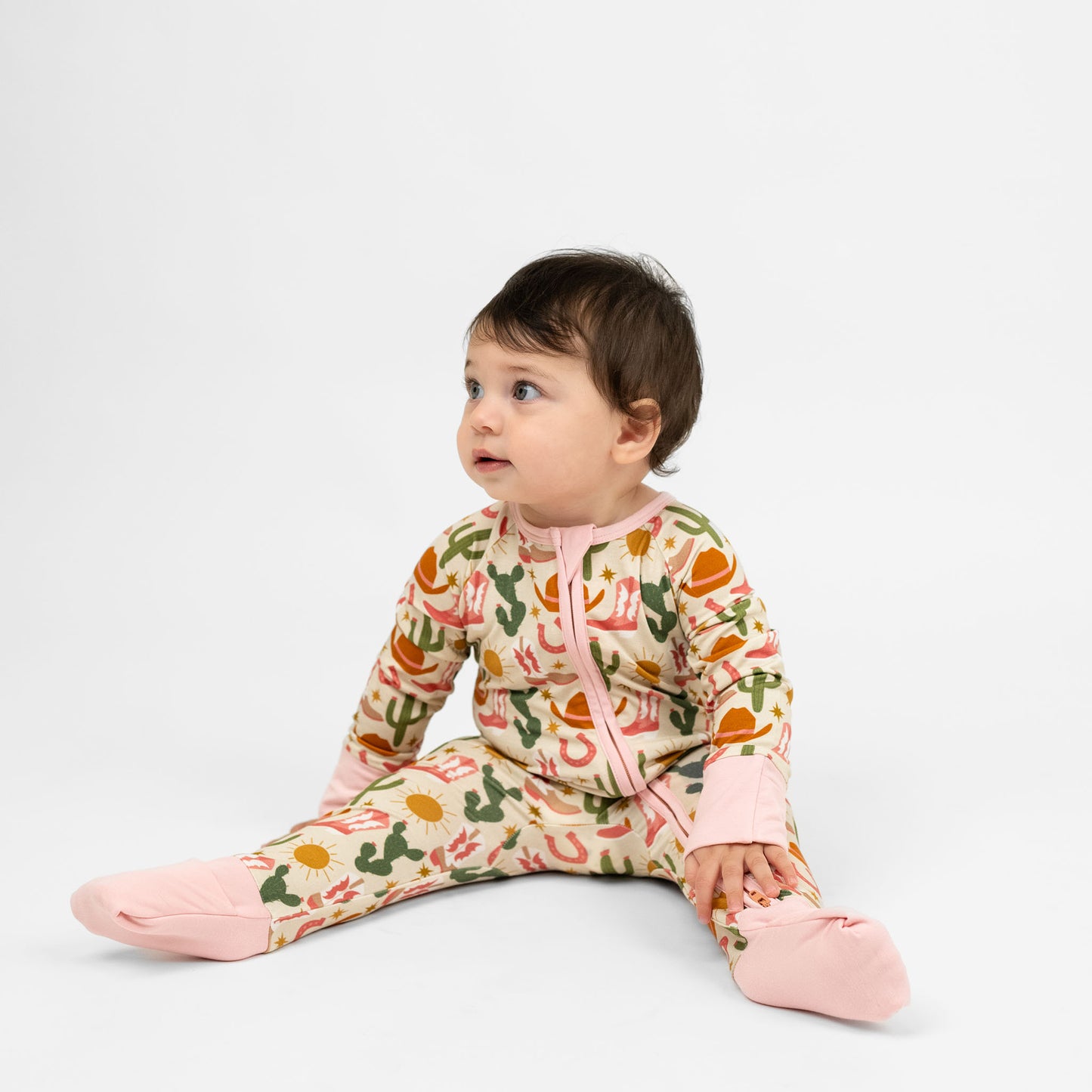 Baby girl wearing our Emerson and Friends Hey Y’all Bamboo Convertible Pajamas. The print shows green cactus, yellow sun, brown cowgirl hat, pink boots and horseshoes. Perfect for cozy rodeo nights or pajama parties with a cowgirl flair!