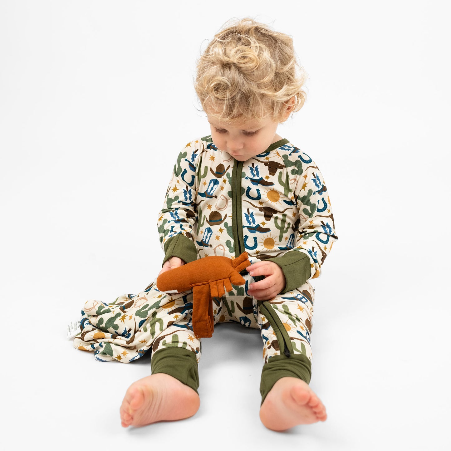 Baby boy holding our lovey featuring a charming cowboy-themed print with blue boots, cacti, suns, horseshoes, and cowboy hats. The perfect blend to pair with our bamboo pajamas.