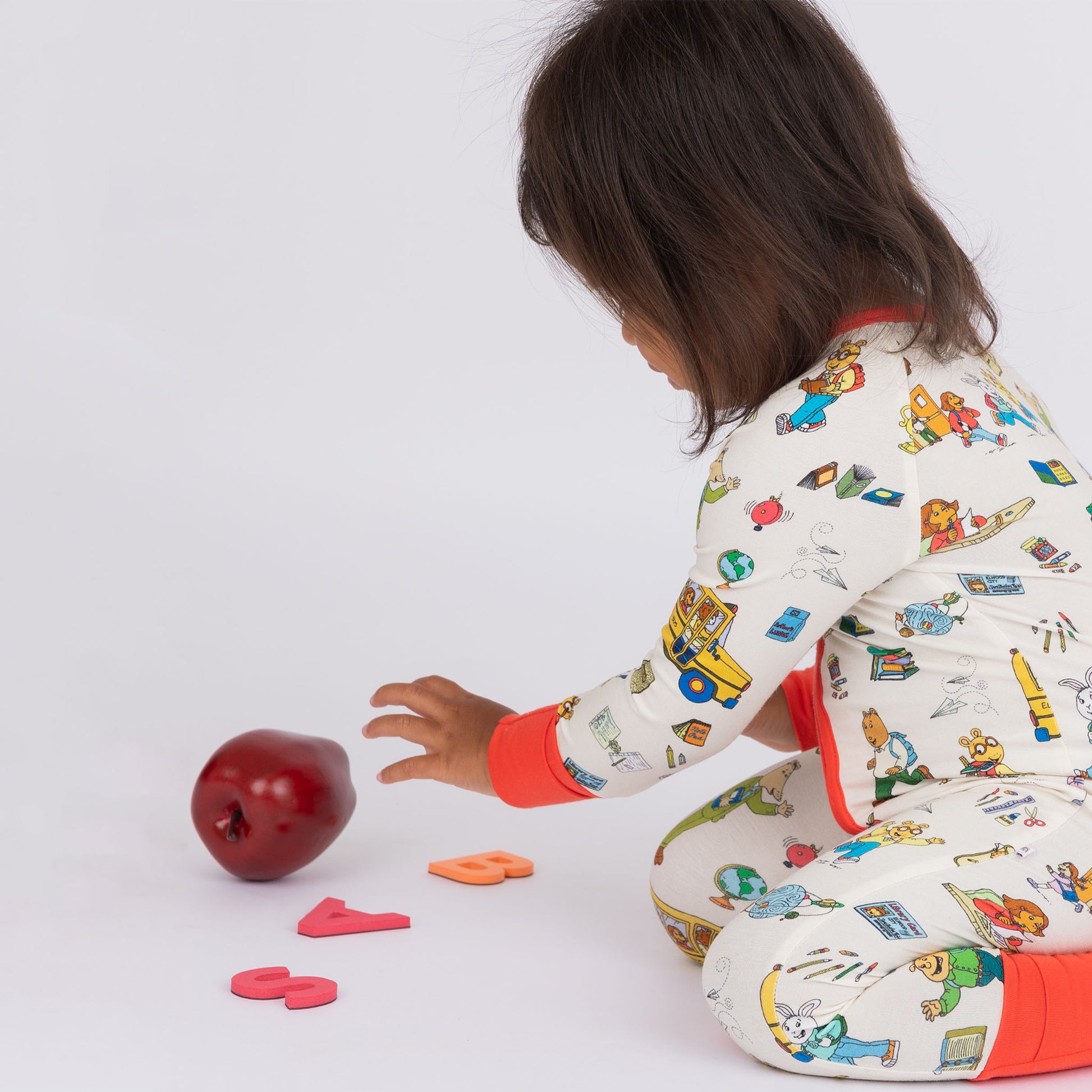 Baby girl sitting in the floor,  wearing our buttery-soft  bamboo convertible pajama featuring red trims and playful illustrations of Arthur™ PBS Character, books, and school bus, a beige background.