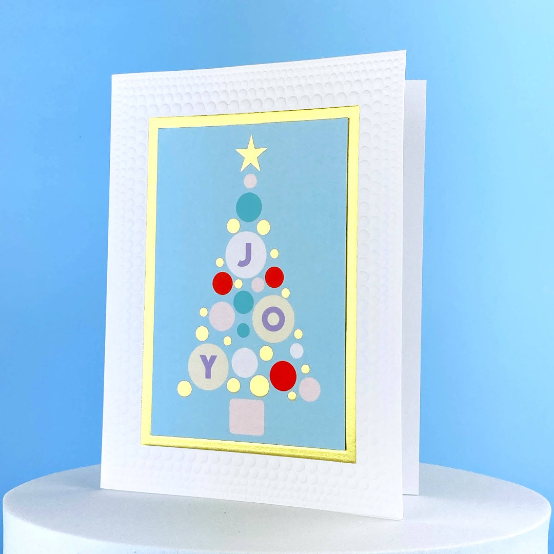 greeting card thats blue and white with a gold trim and a christmas tree made out of dots and the word Joy on it