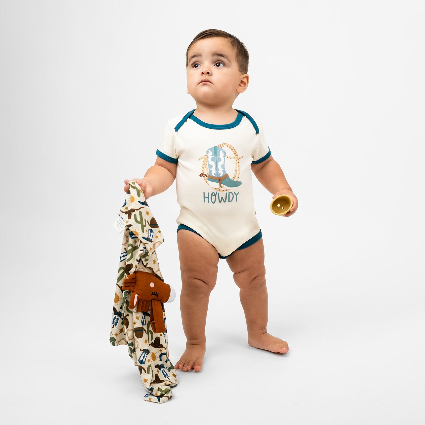 Baby boy holding our lovey featuring a charming cowboy-themed print with blue boots, cacti, suns, horseshoes, and cowboy hats. The perfect blend to pair with our bamboo pajamas.