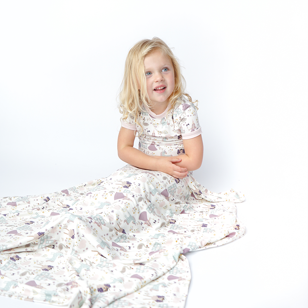 a young girl covers herself with the "once upon a time" bamboo blanket. the "once upon a time" print is a mix of pinks, purples, whites, blues, and yellows. you can see fairy princess and regular princess, unicorns, swans, and castles. theres also hearts, stars and sparkles scattered around the print.