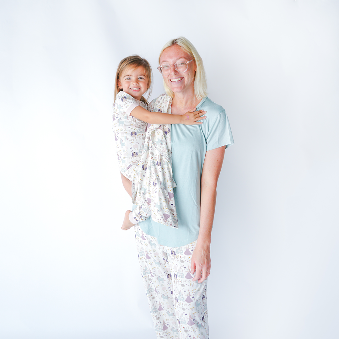 a mom smiles with her daughter on her hip. the "once upon a time" bamboo blanket is draped over her shoulder. they are both wearing "once upon a time" apparel. the mom in the relaxed pants and the daughter in the 2-piece matching pajama set. the "once upon a time" print is a mix of pinks, purples, whites, blues, and yellows. you can see fairy princess and regular princess, unicorns, swans, and castles. theres also hearts, stars and sparkles scattered around the print.