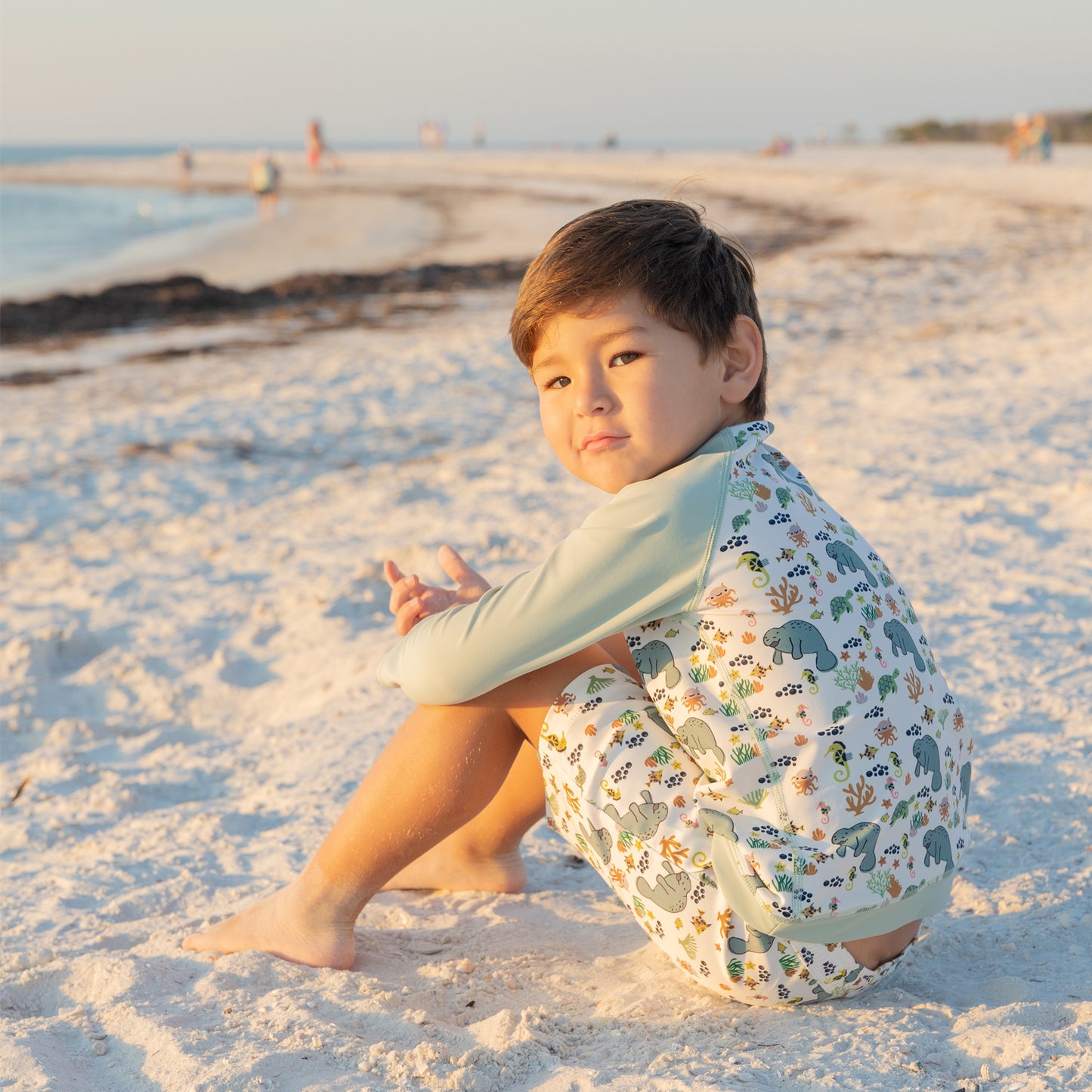 Child sitting on a sandy beach wearing a light blue shirt and patterned shorts.