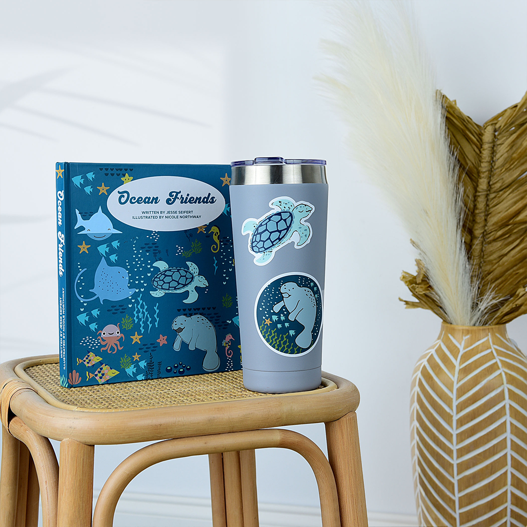 a tervis cup stand on a small table. it has a turtle and the manatee sticker on it. a manatee is centered in the middle of a blue sticker. the manatee is surrounded by sea life that includes starfish, fish, and seaweed. you can also see bubbles. the book "ocean friends" is place behind the cup.