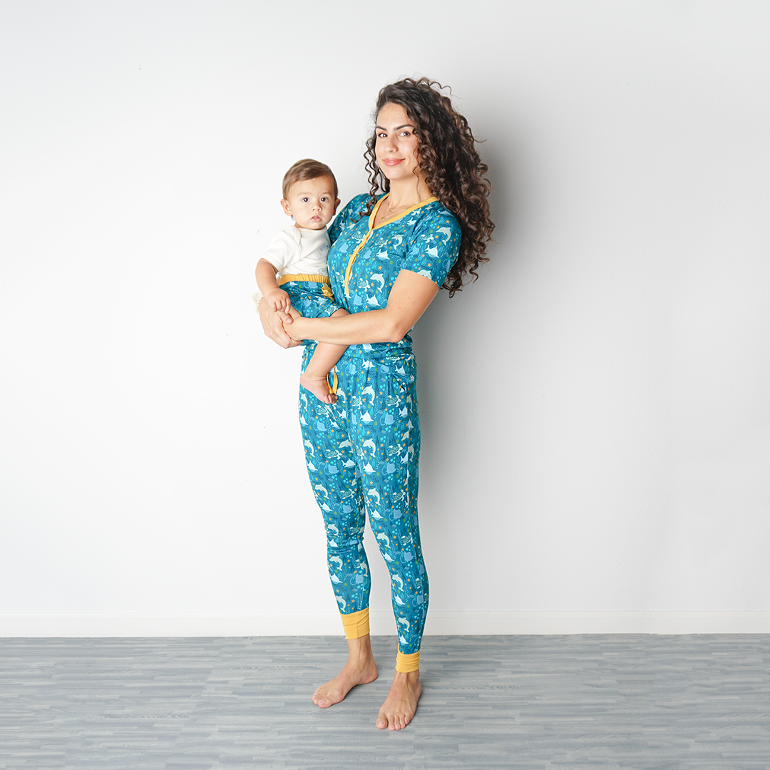 mom stand up against a wall wearing her "ocean friends" women's top and jogger bottoms. she holds her baby on her hip. the "ocean friends" print is a combination of dolphins, stingrays, fish, starfish, coral, bubbles, and sharks, all spread out on a deep sea blue background.