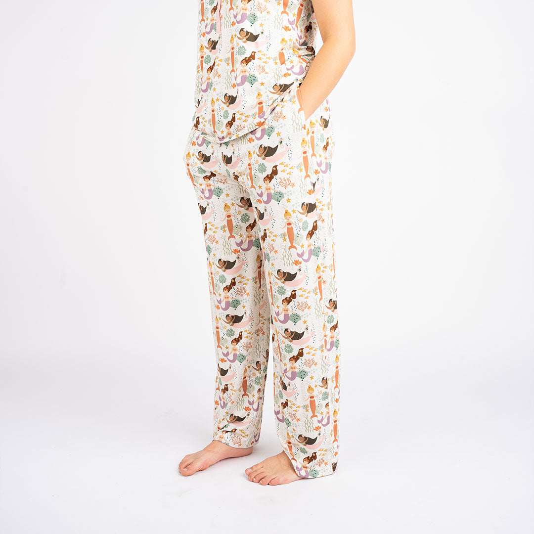 someone poses with their hands in the "making waves" relaxed pants. the "making waves" print has a diverse spread of mermaids, sea coral, starfish, fish, and bubbles all spread out in different colors. this is all put on a beige background.
