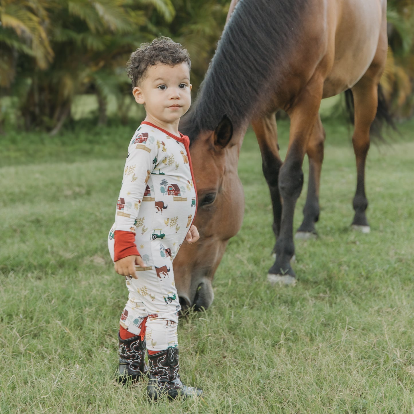 Child wearing On the Farm bamboo convertible pajamas with horse, cow, and tractor print, standing by a horse.