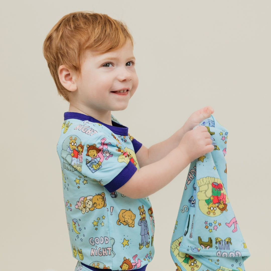 Baby holding our PBS Good Night Arthur™ collaboration blanket with Arthur and friends in a cozy bedtime scene complete with storybooks, toothbrushes, bunny slippers, a sleeping puppy, crescent moons with sleep caps, twinkling stars, and the words "GOOD NIGHT!" on a soft blue background.