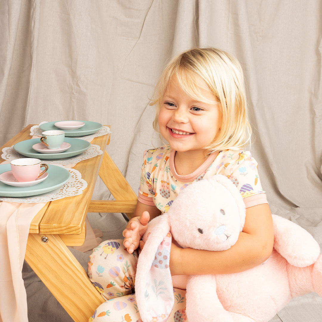 young girl smiles and sits at a table that's set up for a tea party. she holds the pink bunny in her arms