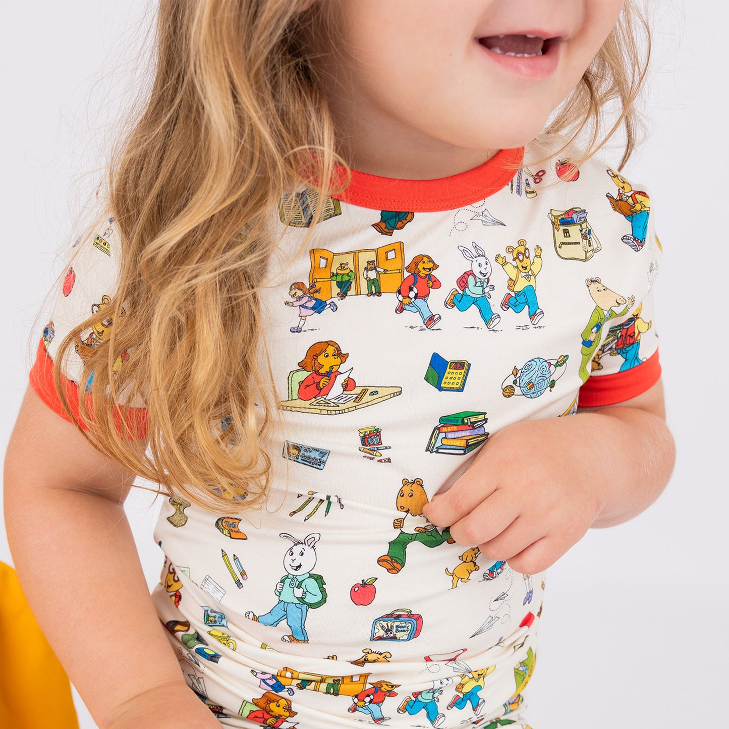 Close up of our short Sleeve Kids Pajama Pants Set from buttery-soft bamboo, featuring red trims and playful illustrations of Arthur™ PBS Character, books, and school bus, a beige background.